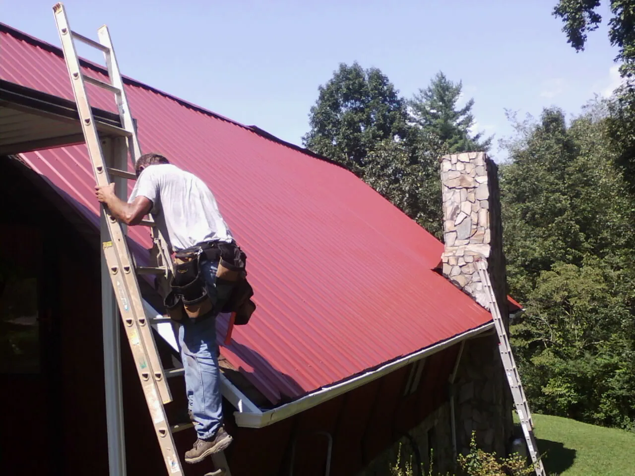 Roofer on a ladder beside a red metal roof and stone chimney — Soffit & Fascia Repair by Roofing Brothers in San Antonio